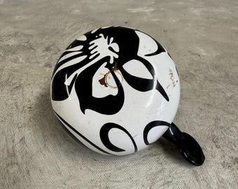 May include: A white bicycle bell with a black and white floral design. The bell has a black handle.