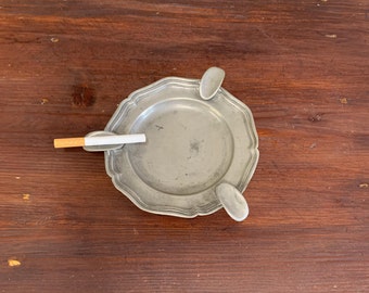 May include: A silver ashtray with three indentations for cigarettes. A cigarette is resting in one of the indentations.