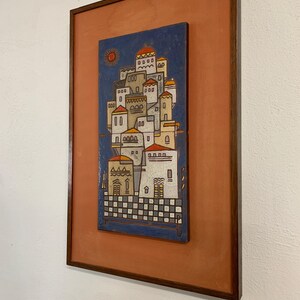 May include: A framed artwork featuring a stylized village scene. The artwork depicts white buildings with red roofs against a blue sky, with a sun in the upper left corner. The frame is a dark brown color, and the background is a muted orange.