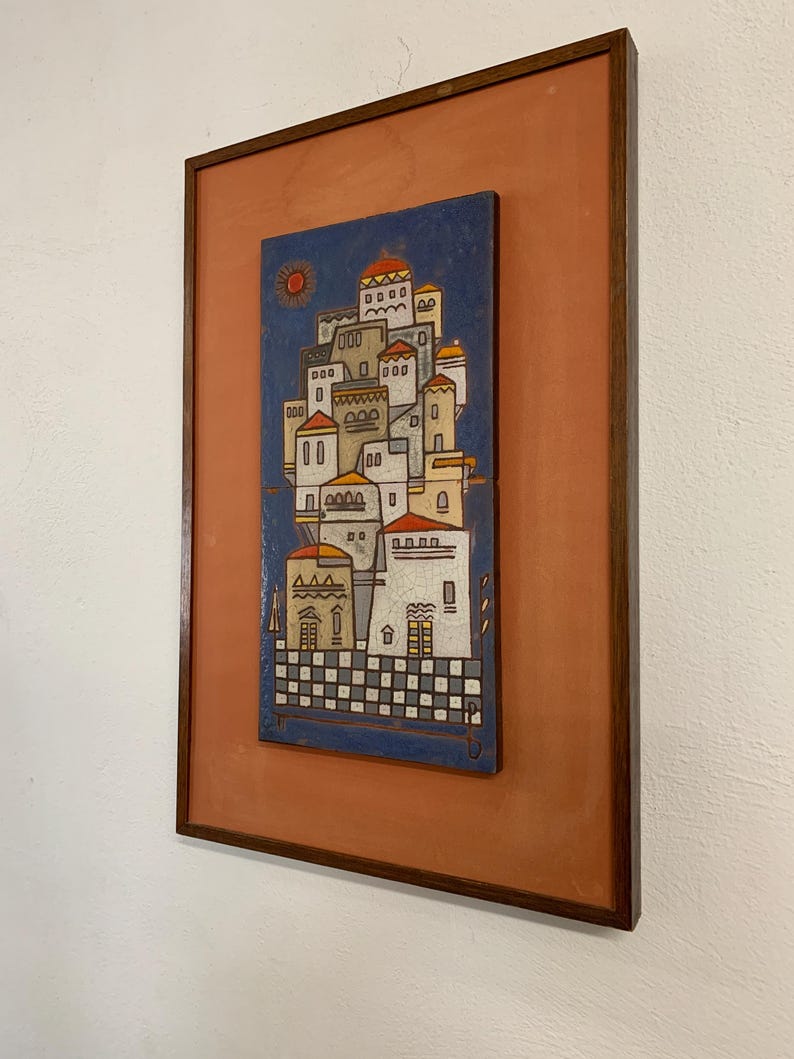 May include: A framed artwork featuring a stylized village scene. The artwork depicts white buildings with red roofs against a blue sky, with a sun in the upper left corner. The frame is a dark brown color, and the background is a muted orange.