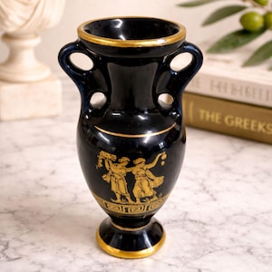 Puede incluir: Un jarrón de cerámica negro con detalles y asas doradas. El jarrón presenta una ilustración dorada de figuras con vestimenta clásica. El borde y la base están adornados con oro. El jarrón mide aproximadamente 15 cm de altura.