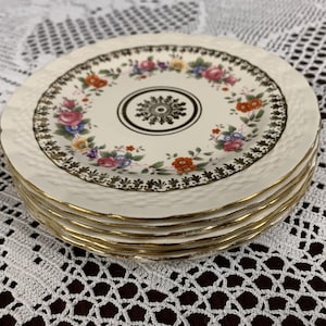 Puede incluir: Un juego de cuatro platos de postre de cerámica blanca con borde dorado y diseño floral. Los platos tienen una superficie texturizada en relieve.