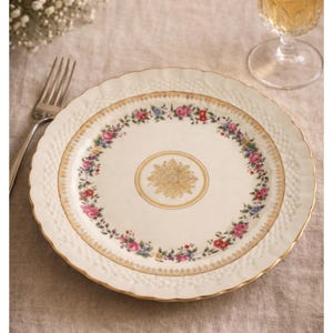 Puede incluir: Plato color crema con diseño floral. El plato presenta un borde dorado y un motivo floral dorado central. Un anillo de flores coloridas rodea el diseño central. El plato tiene un borde texturizado y un borde festoneado.