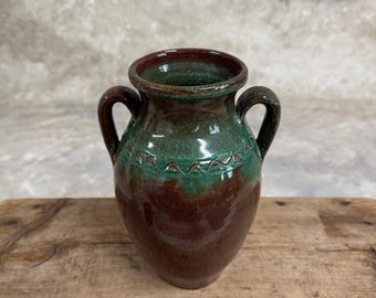May include: A small, green and brown ceramic vase with two handles. The vase has a decorative design around the middle.