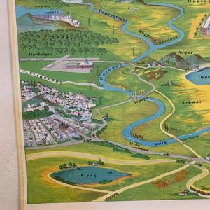 May include: A vintage, illustrated map featuring a river winding through a valley, with mountains in the background. The map includes labels in Greek, depicting a town, lake, and airport. The color palette is dominated by greens, blues, and yellows.