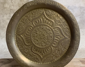 May include: A round, antique brass tray with intricate floral and leaf patterns. The tray's center features a large, symmetrical floral design, surrounded by detailed embellishments. The rim is adorned with a repeating pattern of small dots and floral motifs.