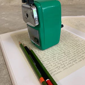 K&ouml;nnte beinhalten: Ein gr&uuml;ner manueller Bleistiftanspitzer mit silbernem Klingengeh&auml;use und schwarzen Akzenten. Drei Bleistifte, gr&uuml;n mit orangefarbenen und roten Spitzen, und ein schwarzer Bleistift liegen auf einem offenen Buch mit handschriftlichem Text.
