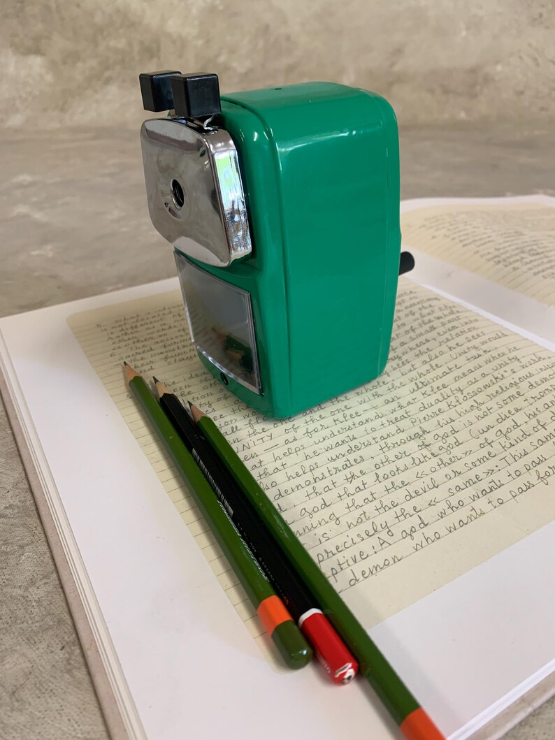 K&ouml;nnte beinhalten: Ein gr&uuml;ner manueller Bleistiftanspitzer mit silbernem Klingengeh&auml;use und schwarzen Akzenten. Drei Bleistifte, gr&uuml;n mit orangefarbenen und roten Spitzen, und ein schwarzer Bleistift liegen auf einem offenen Buch mit handschriftlichem Text.