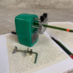 K&ouml;nnte beinhalten: Ein gr&uuml;ner Metall-Bleistiftanspitzer mit einer silbernen Klinge, der einen gr&uuml;nen Bleistift mit einem orangefarbenen Streifen anspitzt. Der Anspitzer liegt auf einem Notizbuch mit handschriftlichem Text. Eine Metallklemme befindet sich im Vordergrund.