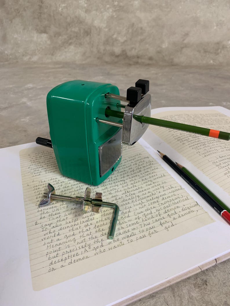 K&ouml;nnte beinhalten: Ein gr&uuml;ner Metall-Bleistiftanspitzer mit einer silbernen Klinge, der einen gr&uuml;nen Bleistift mit einem orangefarbenen Streifen anspitzt. Der Anspitzer liegt auf einem Notizbuch mit handschriftlichem Text. Eine Metallklemme befindet sich im Vordergrund.