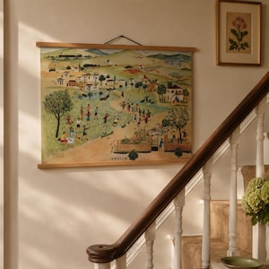 May include: A wall hanging featuring a colorful, detailed illustration of a rural landscape with numerous figures, trees, houses, and a train in the distance. The artwork is mounted between two light-colored wooden bars and suspended by a black cord.