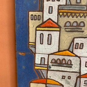May include: A ceramic wall tile featuring a stylized architectural scene with white buildings, orange roofs, and yellow accents against a deep blue background. The surface displays a distinct crackle glaze finish with dark brown outlines.