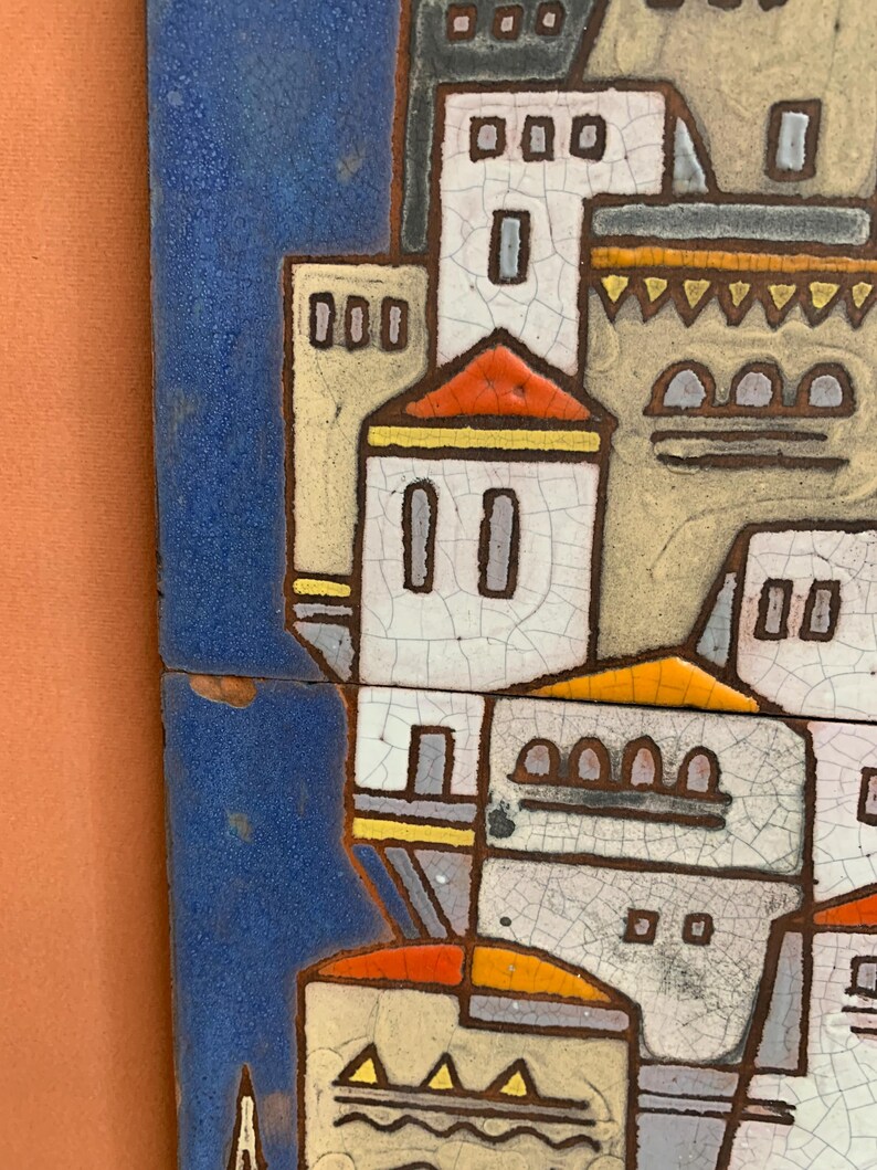 May include: A ceramic wall tile featuring a stylized architectural scene with white buildings, orange roofs, and yellow accents against a deep blue background. The surface displays a distinct crackle glaze finish with dark brown outlines.