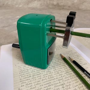 K&ouml;nnte beinhalten: Ein gr&uuml;ner Bleistiftanspitzer mit einer silbernen Klinge spitzt einen gr&uuml;nen Bleistift an. Zwei Bleistifte, einer gr&uuml;n und einer schwarz, liegen auf einer Seite mit handschriftlichen Notizen. Der Anspitzer steht auf einer hellen Oberfl&auml;che.