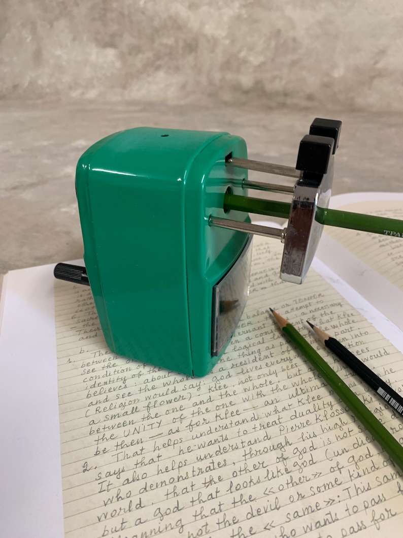 K&ouml;nnte beinhalten: Ein gr&uuml;ner Bleistiftanspitzer mit einer silbernen Klinge spitzt einen gr&uuml;nen Bleistift an. Zwei Bleistifte, einer gr&uuml;n und einer schwarz, liegen auf einer Seite mit handschriftlichen Notizen. Der Anspitzer steht auf einer hellen Oberfl&auml;che.