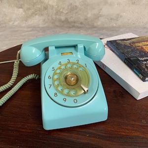 Op de afbeelding: Een retro, turquoise draaischijftelefoon met een doorzichtige, geel getinte wijzerplaat. De telefoon heeft een bijpassende handset en een spiraalsnoer. De telefoon staat op een donker houten oppervlak, met een boek op de achtergrond.