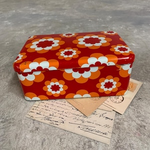May include: A rectangular red metal box decorated with a white and orange floral pattern. The box sits on top of several vintage postcards and letters, featuring handwritten text.