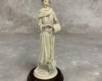 May include: A porcelain figurine of a woman in a long white coat and a hat, holding a leash with a small white dog by her side. The figurine is on a wooden base with the artist's signature.