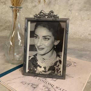 Puede incluir: Un marco de fotos rectangular plateado con un diseño de lazo en la parte superior, que contiene un retrato en blanco y negro de una mujer con un collar de perlas y pendientes. El marco está sobre un libro con texto.
