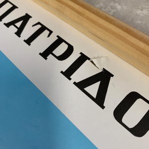 May include: Close-up of a framed print with the Greek word "&Pi;&Alpha;&Tau;&Rho;&Iota;&Delta;&Alpha;" in bold black letters. The print has a light blue background and is framed with natural wood. The paper has a small tear.