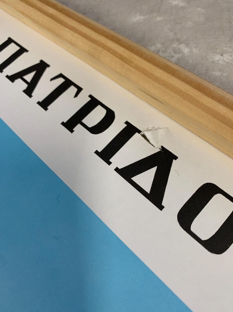 May include: Close-up of a framed print with the Greek word "&Pi;&Alpha;&Tau;&Rho;&Iota;&Delta;&Alpha;" in bold black letters. The print has a light blue background and is framed with natural wood. The paper has a small tear.
