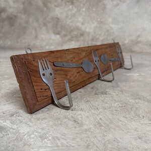 May include: A wooden wall-mounted coat rack with four hooks made from repurposed silverware. The hooks are shaped like forks and spoons.