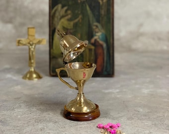 May include: A gold metal incense burner with a bell-shaped lid. The burner is on a wooden base and has a handle. There are pink flower buds and incense on the surface.