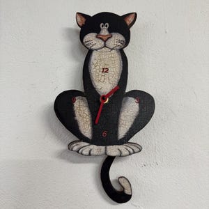 May include: A black and white cat-shaped wall clock. The clock has a black body with white accents and a cracked paint effect. Red clock hands point to the numbers on the clock face. The cat's tail curls upwards.