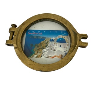 K&ouml;nnte beinhalten: Ein kreisf&ouml;rmiger Bullaugenrahmen in Messingfarbe mit einem Klappdeckel und seitlichen Verschl&uuml;ssen. Der Rahmen enth&auml;lt eine Postkarte mit einer malerischen Ansicht von Santorini, Griechenland, mit wei&szlig;en Geb&auml;uden und blau gekuppelten Kirchen. Das Wort "Santorini" ist sichtbar.