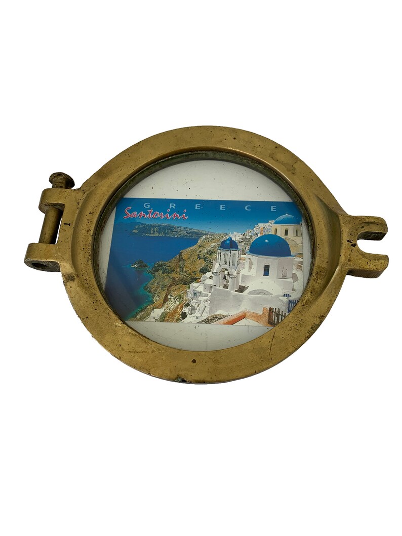 K&ouml;nnte beinhalten: Ein kreisf&ouml;rmiger Bullaugenrahmen in Messingfarbe mit einem Klappdeckel und seitlichen Verschl&uuml;ssen. Der Rahmen enth&auml;lt eine Postkarte mit einer malerischen Ansicht von Santorini, Griechenland, mit wei&szlig;en Geb&auml;uden und blau gekuppelten Kirchen. Das Wort "Santorini" ist sichtbar.
