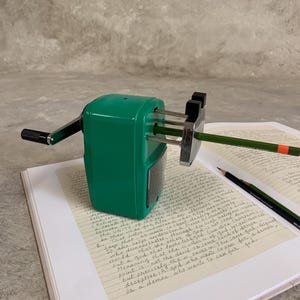 K&ouml;nnte beinhalten: Ein gr&uuml;ner manueller Bleistiftanspitzer mit schwarzem Griff und silbernen Klingen, der einen gr&uuml;nen Bleistift anspitzt. Der Anspitzer steht auf einem Notizbuch mit handschriftlichem Text. Ein zweiter Bleistift liegt daneben.