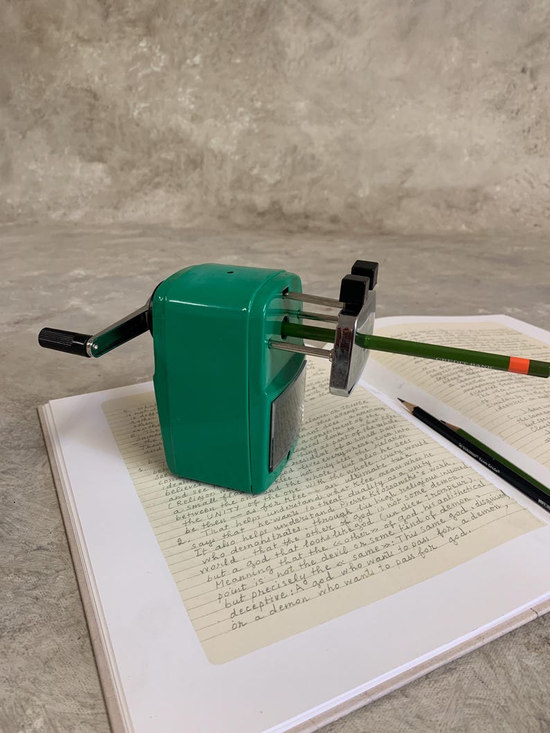 K&ouml;nnte beinhalten: Ein gr&uuml;ner manueller Bleistiftanspitzer mit schwarzem Griff und silbernen Klingen, der einen gr&uuml;nen Bleistift anspitzt. Der Anspitzer steht auf einem Notizbuch mit handschriftlichem Text. Ein zweiter Bleistift liegt daneben.