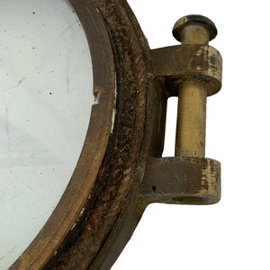 K&ouml;nnte beinhalten: Nahaufnahme eines Vintage-Bullauges aus Messing mit einer klaren Glasscheibe. Das Bullauge hat ein verwittertes, gealtertes Aussehen mit sichtbaren Gebrauchsspuren. Der Rahmen ist dunkelbraun und das Scharnier ist aus Messing.