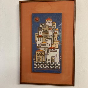 May include: Framed artwork depicting a stylized village scene. The artwork features a blue background with a sun and a cluster of white buildings with red and yellow roofs. The frame is a dark brown color, and the background is orange.