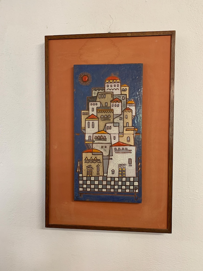May include: Framed artwork depicting a stylized village scene. The artwork features a blue background with a sun and a cluster of white buildings with red and yellow roofs. The frame is a dark brown color, and the background is orange.