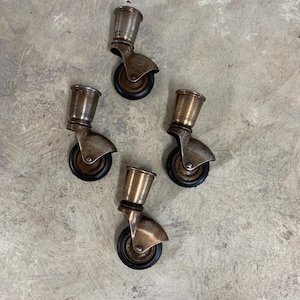 May include: A set of four vintage-style furniture casters featuring metallic brass-toned conical sockets and dark brown rubber wheels. Each caster includes a swivel mechanism and a metal housing, showing signs of age and patina on the metal surfaces.
