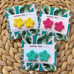 May include: Three pairs of polymer clay earrings in the shape of a flower. The earrings are displayed on cards with the text "esther lian jewellery" and the shop's social media handle and website. The earrings are yellow, pink, and turquoise.