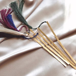 May include: Five gold metal bookmarks with different colored tassels. The tassels are made of yarn and are in the colors of black, white, burgundy, navy blue, and green.