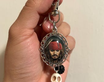 May include: A silver-toned keychain featuring a portrait of a character with a red bandana. The portrait is set in an ornate, oval frame. Below the frame hangs a small, white skull charm and black beads. The keychain is held by a silver clasp.
