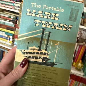 May include: A vintage paperback book titled The Portable Mark Twain with a teal cover featuring a black and white illustration of a steamboat. The text includes the title and a list of included works by the author, with visible wear on the edges.