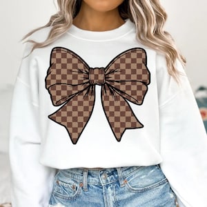 May include: White sweatshirt featuring a large brown bow graphic with a checkered pattern. The bow is outlined in black, creating a bold contrast against the white fabric. The design is centered on the chest.