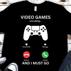 以下が含まれることがあります： 白いビデオゲームコントローラーのグラフィックと「VIDEO GAMES are calling... Remind Me Message Decline Accept AND I MUST GO」というテキストが書かれた黒いTシャツ。