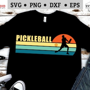Peut inclure: Un t-shirt noir avec un graphique rétro d'une personne jouant au pickleball. Le graphique présente un coucher de soleil avec un schéma de couleurs jaune, vert et bleu. Le texte "Pickleball" est écrit en lettres jaunes au-dessus du graphique.