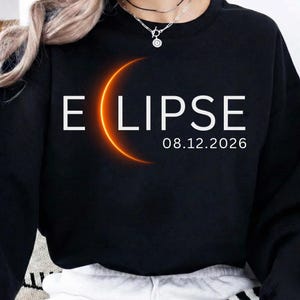 May include: A black long-sleeve sweatshirt featuring the white text "ECLIPSE" with a glowing orange crescent moon replacing the letter "C". Below the text, the date "08.12.2026" is printed in white. The sweatshirt has a crew neckline.