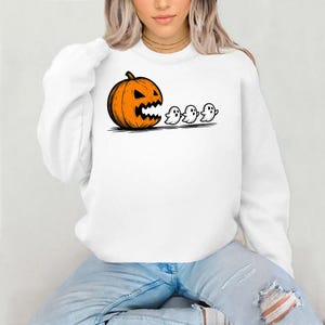 Puede incluir: Sudadera blanca con un diseño de Halloween. El diseño presenta una calabaza naranja con una cara tallada, seguida de tres pequeños fantasmas blancos. El diseño es en negro y naranja.