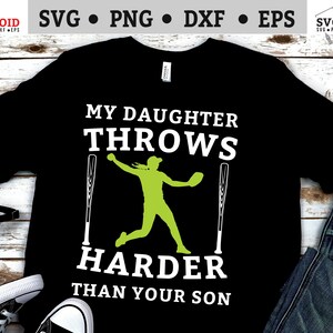 Op de afbeelding: Zwart T-shirt met witte tekst die "Mijn dochter gooit harder dan jouw zoon" luidt. In het midden van de tekst staat een groene silhouet van een softbalspeler.