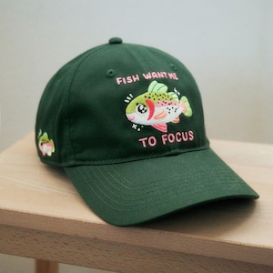 Peut inclure: Casquette de baseball vert foncé avec le texte brodé "FISH WANT ME TO FOCUS" et un dessin de poisson de dessin animé. Le poisson est blanc, rouge, vert et jaune. Un plus petit motif de poisson est sur le côté de la casquette.