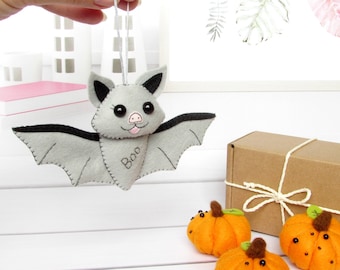 May include: A gray felt bat with black wings and pink nose, embroidered with the word "Boo". The bat is hanging from a string and there are three orange felt pumpkins in the background.