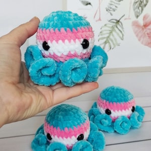 以下が含まれることがあります： 青、ピンク、白の糸で編まれた、3つの編みぐるみタコ。タコは黒い目をしていて、白い表面に並べてあります。