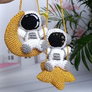 May include: Two crocheted astronaut ornaments hanging from gold chains with gold stars. One astronaut is sitting on a gold crescent moon and the other is sitting on a gold star.